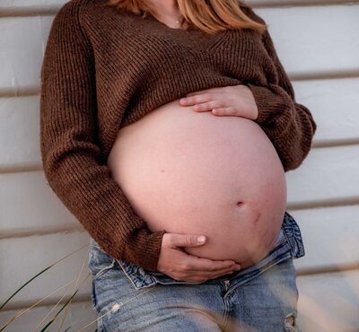 Close up of a pregnant belly with the mother wearing a brown jumper and jeans while leaning against a white weatherboard wall