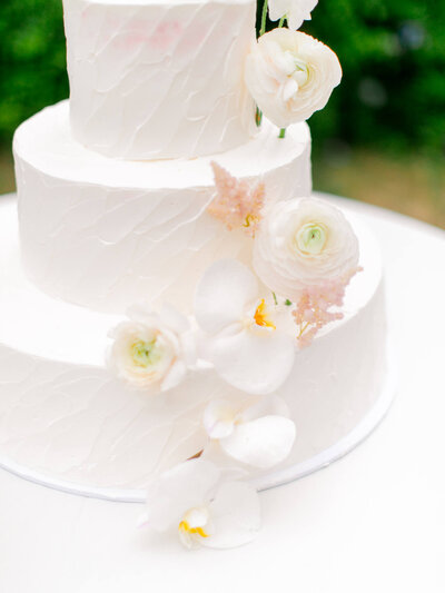 Three-tiered white wedding cake adorned with delicate white flowers, including ranunculus and orchids, on a textured surface. Elegant and minimalistic design.