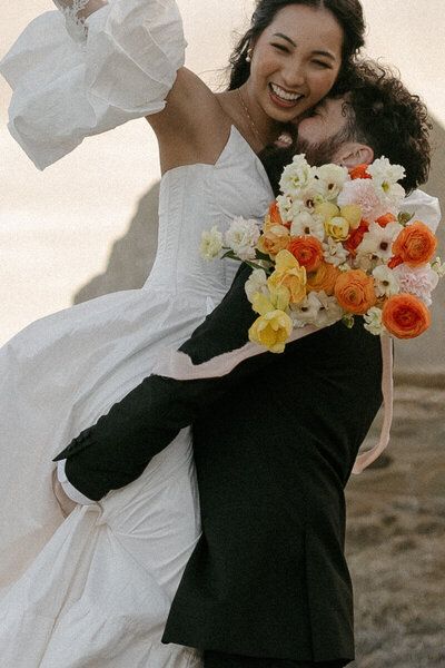 A close-up wedding photo of the bride and her bouquet. The focus of the photo is on the bouquet, and the woman's face is out of the shot. The wedding bouquet is peach, dusty purple, and golden brown spring wedding flowers. The woman is wearing a white dress with sheer sleeves. The setting is the Oregon coast on the beach.