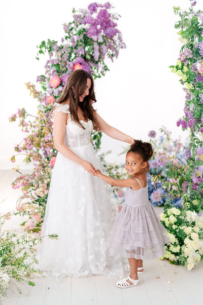 A woman in a white dress holds hands with a young girl in a ruffled lavender dress, both smiling as they stand in front of a vivid floral display of lilacs, roses, and greenery.