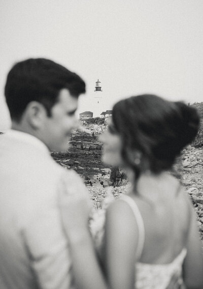 Bride and groom embracing on the rocky Maine coast with a lighthouse in the distance during their wedding day.