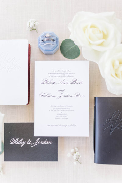 A flat lay design including white roses, eucalyptus, white stationary, black and white vow books, and silver engagement rings and wedding bands by nashville wedding photographer, brooke elliott