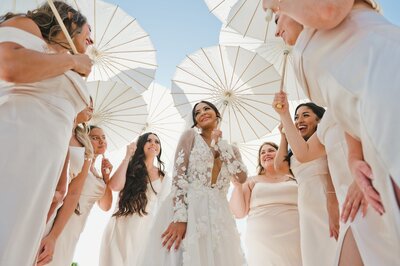 A bride in an elegant white wedding dress in the middle of a group of bridesmaids all holding white umbrellas at a California wedding.