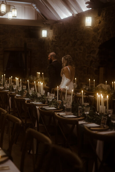 Bride and groom walking away from the camera along a long wedding table set with place settings and tall candles.