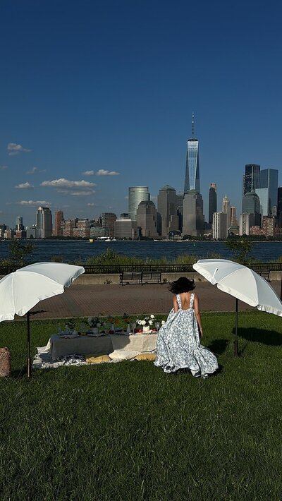 A romantic picnic setup overlooking the New York City skyline, with a woman in a flowing dress seated under a white umbrella, styled by Yours Truly, a New Jersey wedding planner and designer.