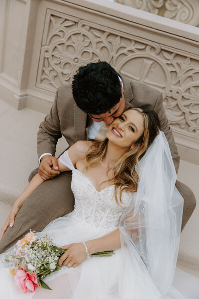 Groom kissing his bride’s cheek as they sit on stone steps, her bouquet and veil flowing over her dress.