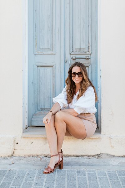 Claire from Lefkas Weddings seated in front of a rustic blue door, wearing sunglasses and a stylish neutral-toned outfit