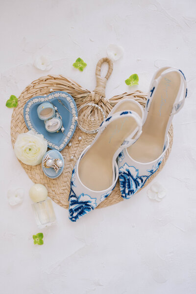 Flat lay of blue and white floral bridal heels styled with ivory roses, jewelry, and perfume on a woven heart mat — elegant details for a refined Southern garden wedding by Harper Mae Events.