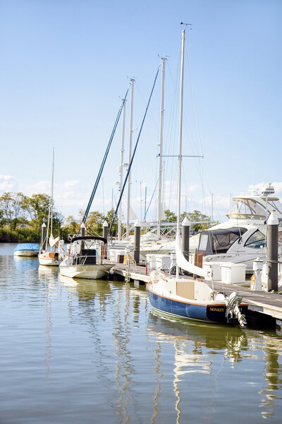 A serene marina scene with several sailboats and yachts docked at a wooden pier. The water is calm, reflecting the boats and the clear blue sky. Trees line the far shore, and a few clouds are visible in the background.