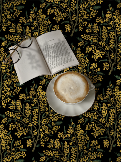 Fabric tablecloth printed with mimosa flowers is shown with an open book, reading glasses, and a frothy cappuccino. Fabric pattern is dark and moody, vintage in mustard yellow, sage green, black.