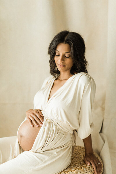 expecting mother sits wearing a cream dress with belly exposed, gently touching her baby bump