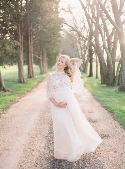 A blonde pregnant mother stands in the middle of a tree-lined lane while the wind blows with one hand under her belly and one behind her head