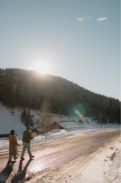 A mom and dad are carrying their baby up a road with a snowy forest behind them.