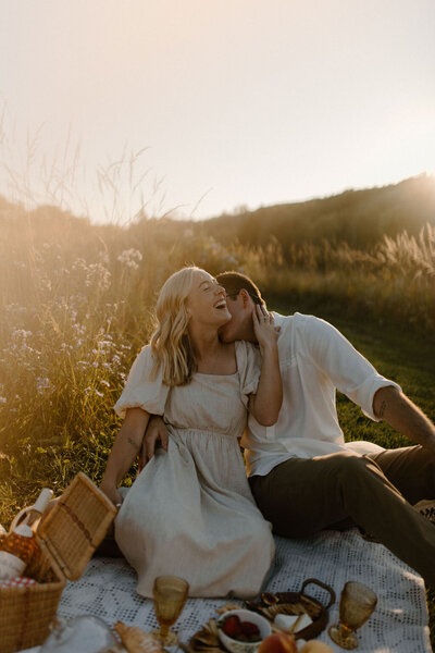Moody, vintage looking golden hour engagement shoot with couple sitting on a picnic blanket in a field with wildflowers and a picnic basket with its contents laid out. The man is kissing the woman on the cheek and she is smiling wide touching his neck. Photo by Morgan Ashley Lynn Photography in Lake Country, WI