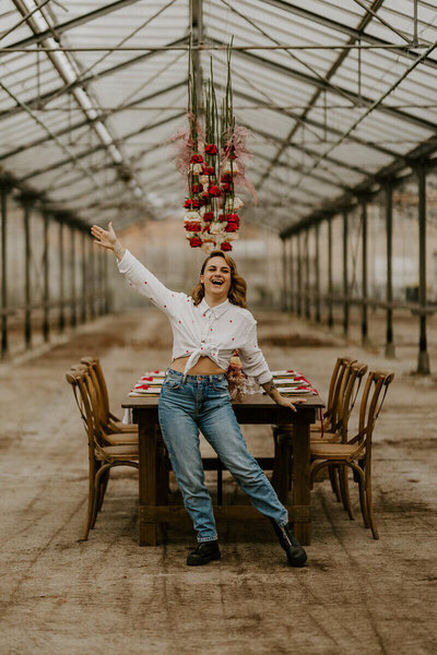 Laura, photographe de mariage en Vendée, pose joyeuse sous un toit de verre décoré de fleurs supendues, appuyée sur une table de mariage décorée pour son workshop photographie de mariage.