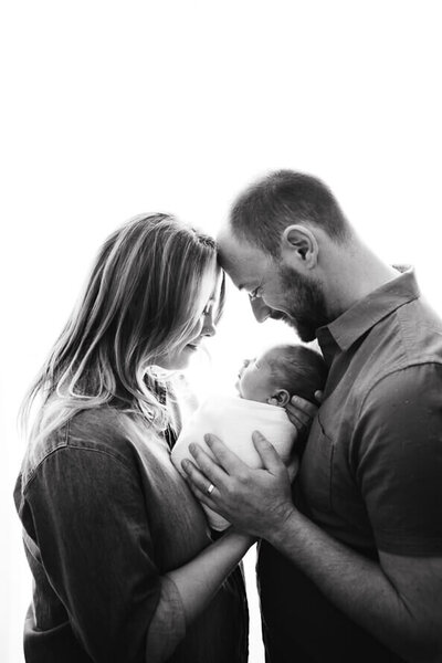 Couple holds their newborn baby wrapped in white with their foreheads together.