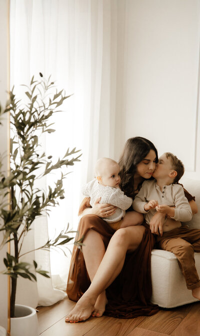 Mother sitting in a softly lit studio holding a baby and receiving a kiss from her older child, capturing a warm and tender family moment in a minimal, earthy-toned setting.