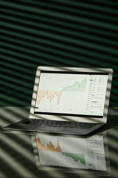 A sleek laptop displaying a financial chart with red and green trend lines, reflected on a shiny surface, set against a textured green background with soft striped shadows.