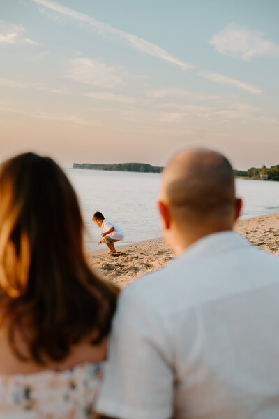 Family photo of mom and dad looking at their son playing on the beach in Kent Island Maryland.