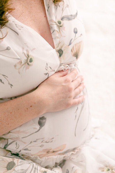 Close up of woman's pregnant belly with her hand resting on top of her belly. She is wearing a light colored floral dress. Light and airy maternity portrait studio in Portland Oregon.