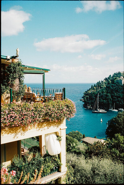 A coastal view featuring a building adorned with colorful flowers and a balcony. Below, a vibrant blue sea stretches towards tree-covered cliffs, with sailboats calmly anchored in the water against a clear, sunny sky.