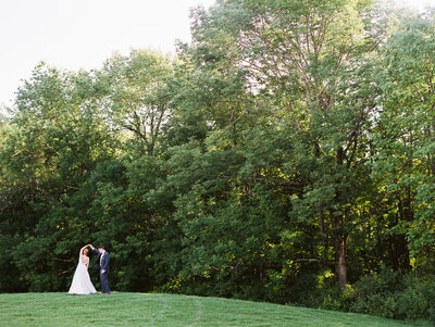 Far away photo of a bride and groom on a grassy landscape with trees behind them