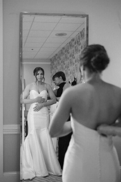 Bride looks at her reflection in the mirror while mom buttons mermaid style wedding gown