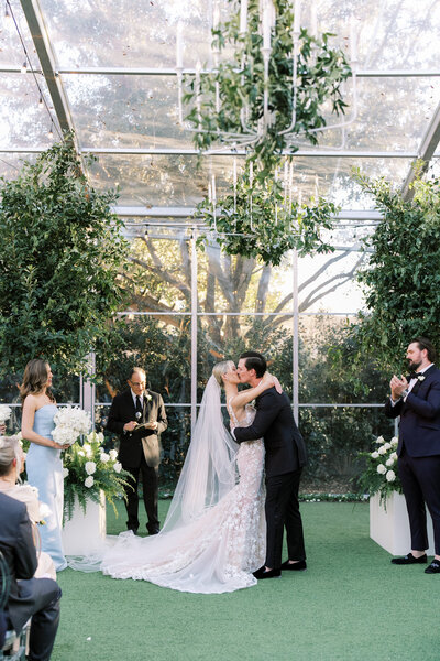 Bride and groom first kiss at their tented wedding in Dallas, with chandeliers covered in greenery