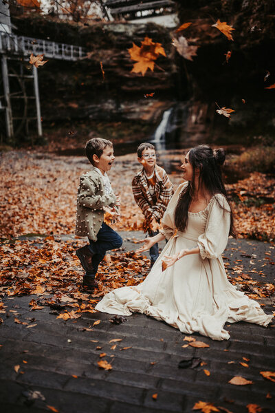 Mother and two sons throw leaves up in the air in front of small waterfall and water mill in Lancaster Ohio