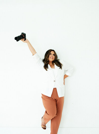 Adriana of Adriana Montoya Photo holding her right arm above her head while holding her camera. Her right leg is also slightly bent and raised off of the ground. She is looking  slightly upwards and to the side, smiling.