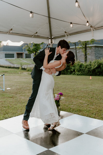 A newlywed couple shares a romantic kiss during their first dance under a tent. They stand on a black and white checkered floor with soft, warm lighting.