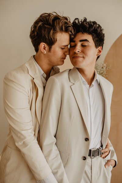 Male couple hold each other closely  in neutral color suits on their wedding day