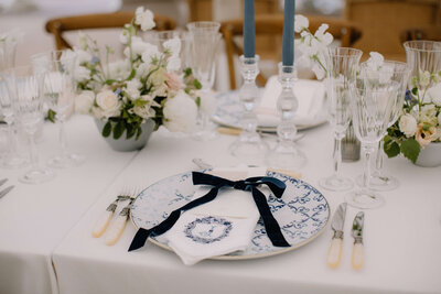 Elegant wedding table setting featuring fine china with blue patterns, velvet ribbon-tied menus, and crystal glassware, complemented by soft white floral centerpieces.