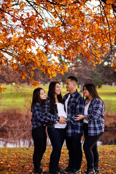 A pregnant woman, her husband and two children are standing under a tree with orange leaves and looking at eachother.