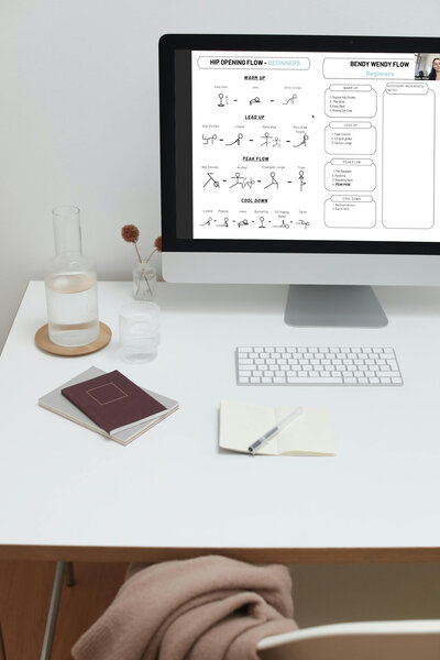 A workspace with a computer monitor displaying 'Hip Opening Flow - Beginners' and 'Bendy Wendy Flow' yoga sequences. The desk is organized, with a notebook, pen, decorative carafe and glass, hinting at a tranquil, productive environment for planning or participating in a yoga class.