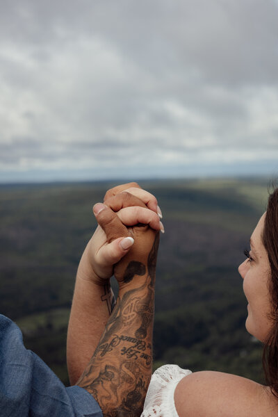 bride and groom interlock hands and are looking at each other while facing the edge of a cliff overlooking nys mountain ranges