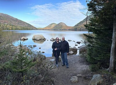 A man and a woman, holding hiking poles, are standing in front of Jordan's Pond at Acadia National Park with