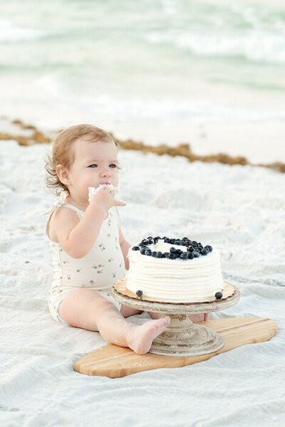 Toddler enjoys a blueberry cake smash on the beach, sitting barefoot on a blanket with soft ocean waves in the background.