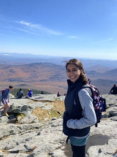 A person with a backpack smiles while standing on a rocky mountaintop, captured beautifully by a New England Family Photographer. The landscape reveals rolling hills adorned in autumn foliage under a clear blue sky, with fellow hikers dotting the background.