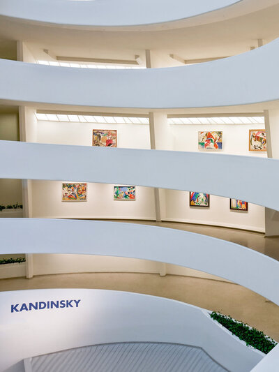 Interior view of an art gallery with white, curving walls and abstract paintings displayed in alcoves. The name Kandinsky is visible on one wall. The architecture features sleek, modern lines and a bright, minimalistic design.