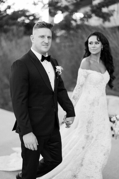 Groom looking into the camera while walking hand in hand with the bride in a classic black and white wedding portrait