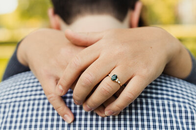 a macro photograph of a woman's hands around her partner's shoulders highlughting her gold engagement ring with a blue oval jewel