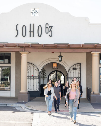 A group of people walks out of SoHo63, a Chandler wedding and event venue, under a sunny sky. The venue’s distinctive white stucco exterior and arched wrought-iron doors are visible behind them, showcasing the stylish entrance of SoHo63.