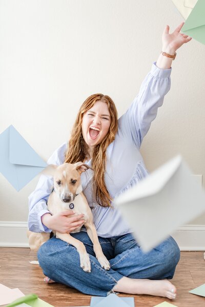 Elyse with her dog Champ in her lap, tossing envelopes in different colors in the air while celebrating.