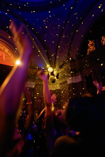 A crowd of people dance under a ceiling with decorative arches. Specks of light illuminate the scene, creating a lively atmosphere.