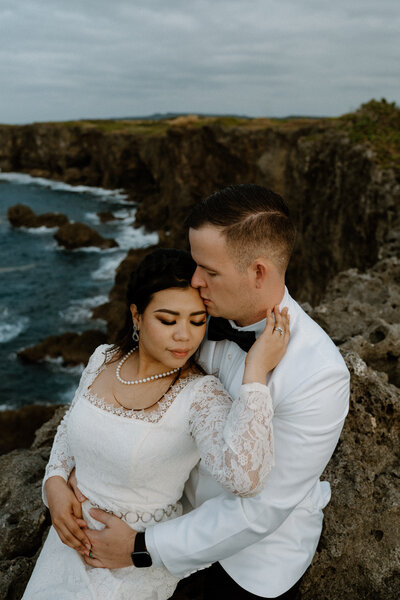 M sitting in front of J, as J kisses M's forehead, M holding her hand against J's neck, while their other hands hold hands, with the oceanside cliffs behind them