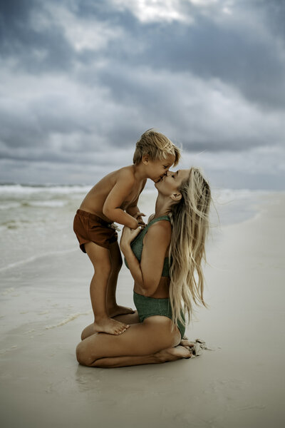 Family Photographer, a mother gives her young son a kiss as he stands on her knees as she kneels near the ocean, they are in their swimsuits