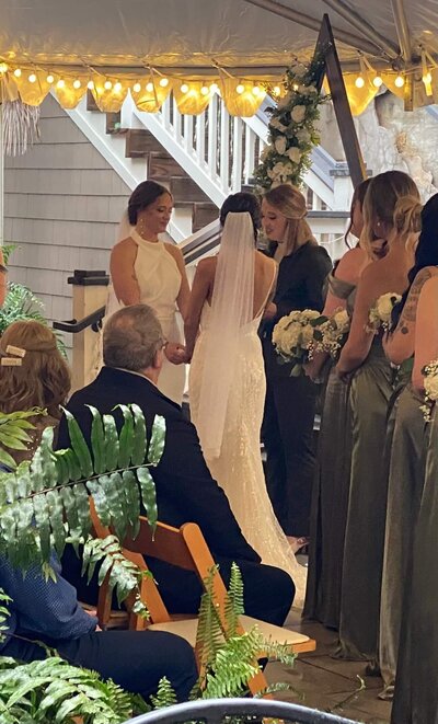 Two brides stand holding hands during their wedding ceremony under a tent, surrounded by bridesmaids in green dresses and guests seated on wooden chairs. The scene is decorated with greenery and string lights.