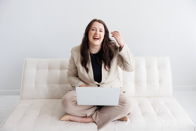 Woman sitting cross-legged on a modern white couch, working on a laptop – professional brand and web designer helping women entrepreneurs build bold brands