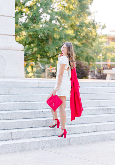 A person stands on stone steps in front of a large stone building, holding a bright red coat. They are wearing a white dress and red shoes. Trees are visible in the background.